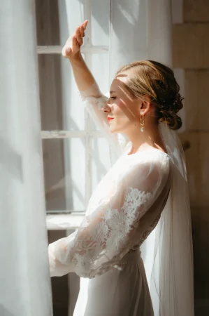 Une mariée avec sa robe posant devant une fenêtre en pleine lumière sur son lieu de mariage
